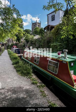 Hell gestrichene schmale Boote auf dem Springs Branch des Leeds-Liverpool Canal, die entlang des Treidelpfades liegen und von einer Kirche und einem Pub überblickt werden. Stockfoto