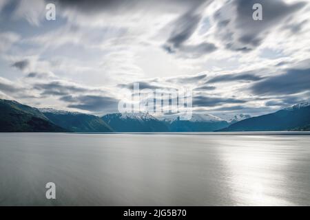 Blick über einen Teil des Südfjords auf teilweise schneebedeckte Berge, die Sonne glitzert im Wasser, lange Belichtung, wodurch das Wasser glatt aussieht Stockfoto