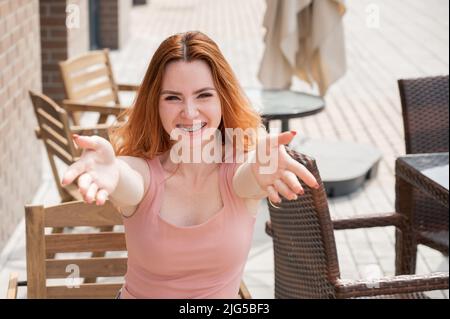 Die junge rothaarige Frau sitzt in einem Straßencafé und streckt ihre Arme zur Umarmung. Stockfoto