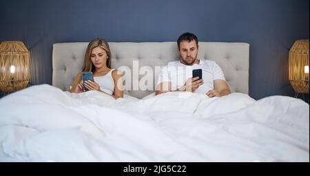 ID eher scrollen als rufen. Aufnahme eines jungen Paares, das nach einem Streit im Bett seine Telefone separat benutzt. Stockfoto