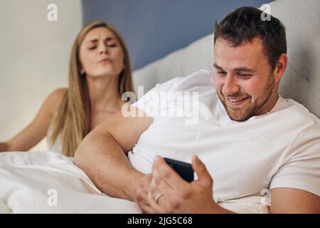Es wird nie enden. Aufnahme einer Frau, die über die Schulter ihres Mannes schaut, während er jemand anderen im Bett textete. Stockfoto