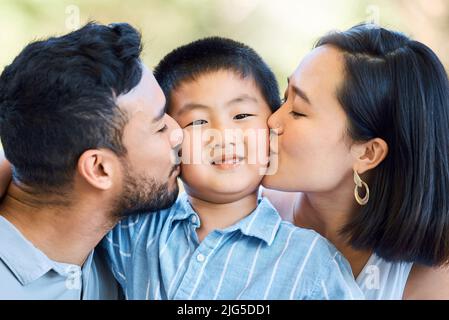 Halten Sie die Küsse kommen. Aufnahme eines entzückenden kleinen Jungen, der von seinen Eltern in einem Garten geküsst wird. Stockfoto