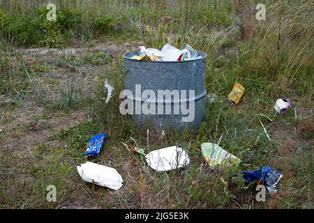 Überfüllte Mülltonne auf einer Wiese an sonnigen Sommertagen. Mülleimer in der Natur. Stockfoto