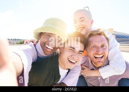 Huckepack-Freunde nehmen an einem sonnigen Tag zusammen ein lächelndes Selfie im Freien auf. Selbstporträt einer Gruppe junger Menschen, die Spaß miteinander haben. Jungs und Mädchen Stockfoto