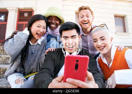 Gruppe lächelnder Menschen, die im Freien ein Mobiltelefon benutzen. Fröhliche Jungs und Mädchen Freunde, die Spaß haben, etwas am digitalen Display zu sehen. Frohe Ferien Stockfoto