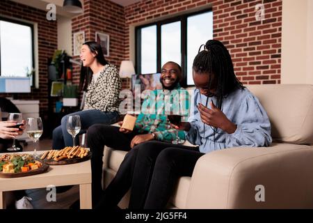 Fröhliche, vielfältige Menschen lachen zusammen auf einer Weinparty im Wohnzimmer zu Hause. Eine multiethnische Gruppe von Freunden lächelt herzlich, während sie die Geburtstagsfeier mit alkoholischen Getränken und Snacks feiert. Stockfoto