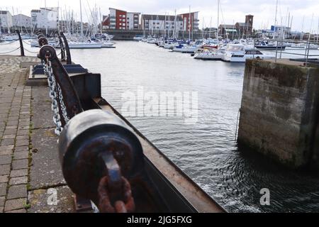 Eine rostige Kette sichert einen Betonpfoller mit Booten, die im Hintergrund an einem modernen Yachthafen angedockt sind Stockfoto