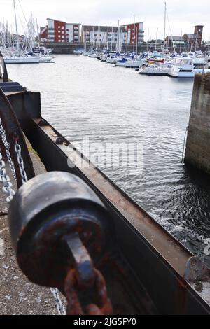 Eine rostige Kette sichert einen Betonpfoller mit Booten, die im Hintergrund an einem modernen Yachthafen angedockt sind Stockfoto