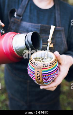 Nicht erkennbare Frau, die ihren Partner mit einer roten Thermo in einem bunten ethnischen Kumpel bedient. Vertikal Stockfoto