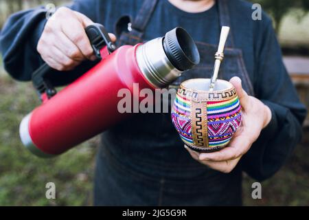 Nicht erkennbare Frau, die ihren Partner mit einer roten Thermo in einem bunten ethnischen Kumpel bedient. Stockfoto