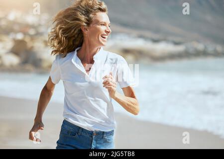 Fröhliche reife Frau, die an einem sonnigen Tag am Strand läuft. Schöne Frau mittleren Alters, die lacht, aktiv ist und Spaß hat während der Sommerferien Stockfoto