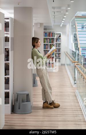 In voller Länge nachdenkliche Leserin mittleren Alters mit Buch in der öffentlichen Bibliothek Stockfoto