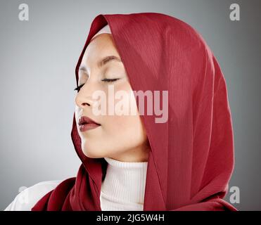 Stilvolle junge muslimische Frau in einem roten Hijab mit geschlossenen Augen auf einem grauen Studiohintergrund. Frau aus dem Nahen Osten trägt Make-up und ein Kopftuch Stockfoto