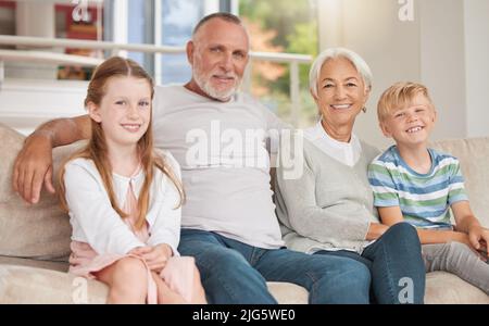 Zwei Geschwister entspannen sich mit ihren Großeltern zu Hause auf einer Couch. Schwester und Bruder besuchen Großeltern. Ein glückliches kaukasisches reifes Paar Stockfoto