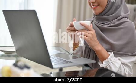 Zugeschnittenes Bild, attraktive asiatische muslimische Frau, die morgens Kaffee oder Tee schlürft, während sie im Wohnzimmer einen Laptop benutzt. Stockfoto