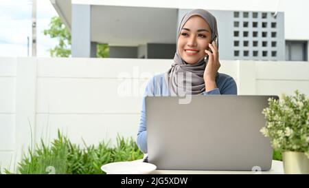 Schöne tausendjährige asiatische muslimische Frau oder Geschäftsfrau, die in ihrem Hinterhof sitzt, am Laptop arbeitet und mit jemandem am Telefon spricht Stockfoto