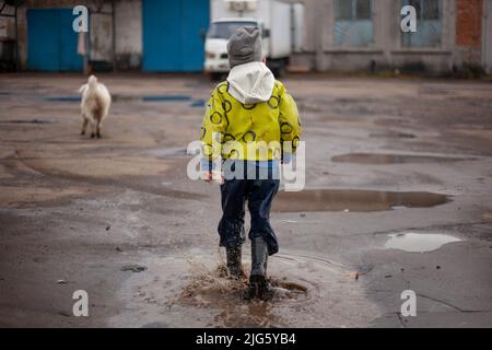 Ein Kind auf einem Spaziergang. Der Typ in der hellen Jacke hat Spaß auf der Straße. Lustiger kleiner Junge. Gummistiefel an den Füßen des Kindes. Stockfoto