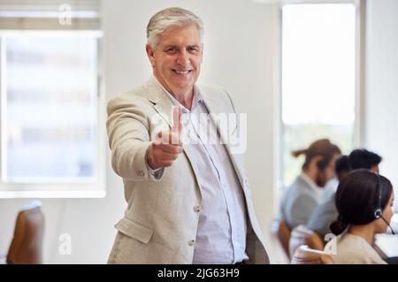 Überlasst die harte Arbeit meinem Team. Aufnahme eines reifen Geschäftsmannes, der im Büro steht und den Daumen nach oben zeigt. Stockfoto