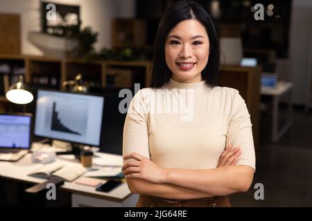 Porträt einer lächelnden asiatischen selbstbewussten Geschäftsfrau mit gekreuzten Armen, die nachts im Büro steht Stockfoto