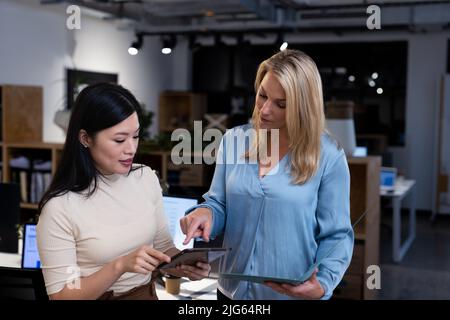 Kaukasische Geschäftsfrau, die der asiatischen Kollegin über ein digitales Tablet in der Nacht im Büro erklärt Stockfoto