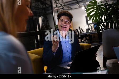 Lächelnder asiatischer Geschäftsmann, der über einen Laptop arbeitet und mit einer kaukasischen Kollegin im Büro spricht Stockfoto