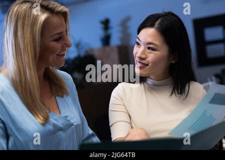 Glückliche, multirassische weibliche Mitarbeiter, die sich gegenseitig anguckend betrachten, während sie den Finanzbericht im Amt analysieren Stockfoto
