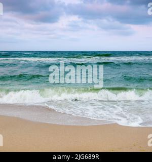 Sandstrand vor dem Sturm. Abendlandschaft mit schweren Wolken in buntem Licht. Türkisfarbene Wellen rauschen am Ufer. Windiges Wetter Stockfoto
