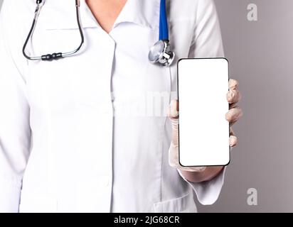 Arzt hält Telefon mit Bildschirm-Mockup. Online-Medizin, mit medizinischen Anwendung im Smartphone, telemedizinische Konzept. Frau im Labormantel mit Stethoskop und Handschuhen. Hochwertige Fotos Stockfoto