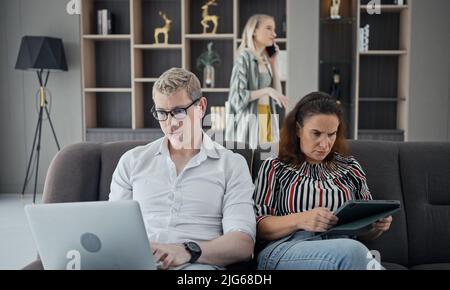 Sucht nach Gadgets. Junge Kaukasierin von drei hält und mit verschiedenen elektronischen Geräten, während sie auf dem Sofa im Wohnzimmer zu Hause sitzt. Eltern und Stockfoto