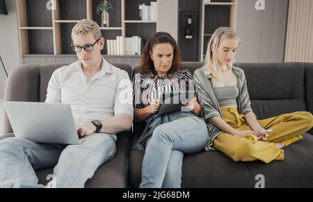 Sucht nach Gadgets. Junge Kaukasierin von drei hält und mit verschiedenen elektronischen Geräten, während sie auf dem Sofa im Wohnzimmer zu Hause sitzt. Eltern und Stockfoto