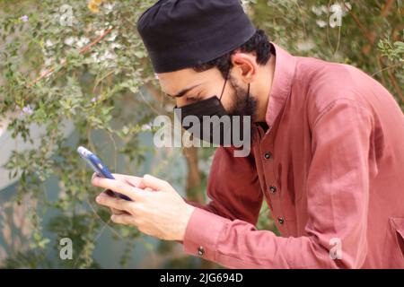 Ein Asain-Typ mit Mütze und schwarzer Maske im Shalwar-Kameez, der mit dem Smartphone im Garten Blumen und Pflanzen fotografiert Stockfoto