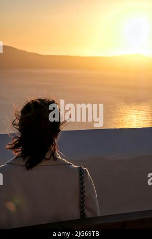 Santorini, Griechenland - 14. Mai 2021 : Blick auf eine Frau, die auf einer Bank sitzt und einen spektakulären goldenen Sonnenuntergang in Fira Santorini sieht Stockfoto