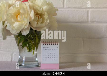 Kalender für Juli 2022 und Pfingstrosen auf dem Tisch. Desktop-Kalender für die Planung Stockfoto