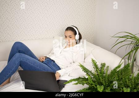 Ein junges, schönes Mädchen liegt auf einem Bett mit Computer und Kopfhörern, in Jeans und weißer Bluse. Stockfoto
