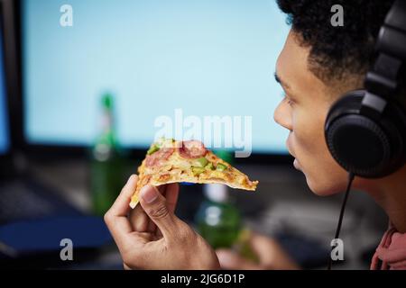 Essen, schlafen, spielen, wiederholen. Nahaufnahme eines jungen Mannes, der Pizza isst, während er zu Hause Videospiele spielt. Stockfoto