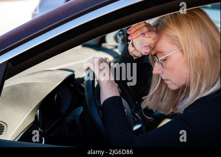 Nachdenkliche Blondine mittleren Alters in Brille sitzt hinter dem Steuer eines Autos, Blick durch das Fenster Stockfoto