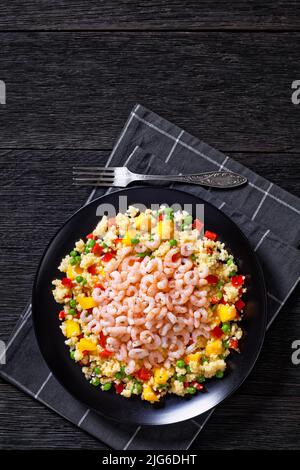 Gekochte kleine rosafarbene Garnelen auf Hirse-Pilaw-grünen Erbsen, roten Paprika und Mangostückchen auf schwarzem Teller auf dunklem Tisch, vertikale Ansicht von oben Stockfoto