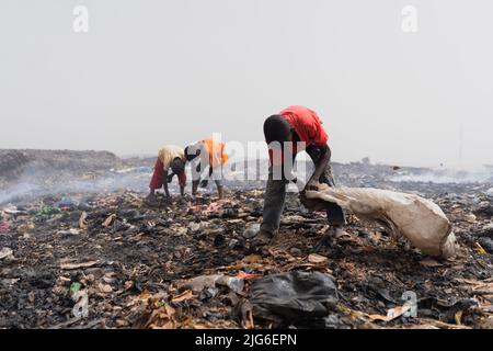 Drei kleine afrikanische Kinder, die auf der Müllhalde nach recycelbaren Wertsachen suchen, inmitten von Abfallhaufen, Schmutz und schwelenden Rauch Stockfoto