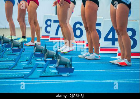 Leichtathletik, Laufen und Sprinten. Beine von Profisportlern vor dem Start eines Sprint-Rennens. Weibliche Athleten stehen vor der blauen Piste Stockfoto