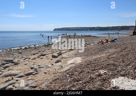 AXMOUTH, DEVON, ENGLAND - JULI 12. 2020: An einem schönen Sommertag paddeln die Menschen im Meer am Axmouth Kiesstrand Stockfoto