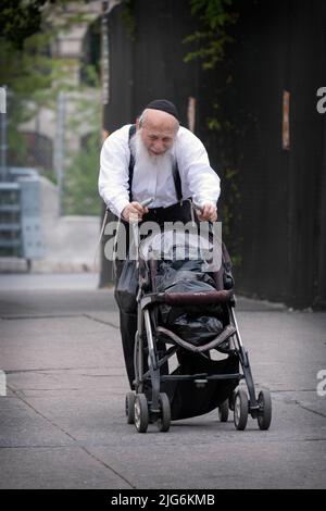 Ein orthodoxer jüdischer Mann, wahrscheinlich ein Rabbi, geht mit Schwierigkeiten mit einer Kutsche als Spaziergänger und um seine Pakete zu tragen. An der Bedford Ave in Brooklyn, NYC. Stockfoto