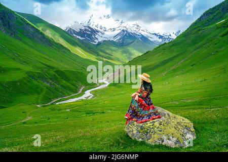 Tourist sitzt auf dem Felsen auf der grünen Weide gegen den höchsten georgischen Berg Shkhara in der Nähe von Ushguli in Georgien. Stockfoto