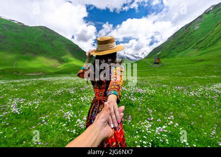 Weibliche Touristen halten die Hand des Mannes und führen ihn auf die grüne Weide gegen den höchsten georgischen Berg Shkhara in der Nähe von Ushguli in Georgien. Stockfoto