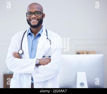 Der freundlichste Arzt der Gegend. Aufnahme eines jungen männlichen Arztes, der mit gekreuzten Armen in einem Krankenhaus in einem Büro stand. Stockfoto