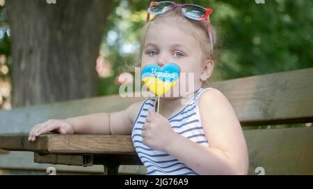 Das kleine blonde Mädchen hält Lebkuchen in ukrainischen Nationalfarben in der Hand, es sagt, die Ukraine sei ich. Nahaufnahme des Mädchens, das auf der Parkbank sitzt Stockfoto