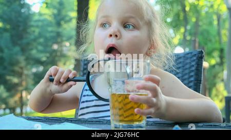 Kleines Mädchen schaut in die Linse um. Nahaufnahme eines blonden Mädchens, das die Welt um sie herum studiert und sie durch eine Lupe betrachtet, während es im Raum sitzt Stockfoto