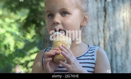 Nettes kleines Mädchen isst draußen Eis. Nahaufnahme eines blonden Mädchens, das auf der Parkbank sitzt und Eis isst. Odessa, Ukraine Stockfoto