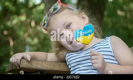 Das kleine blonde Mädchen hält Lebkuchen in ukrainischen Nationalfarben in der Hand und sagt: „Die Ukraine ist ich“. Nahaufnahme des Mädchens, das auf dem Park sitzt Stockfoto