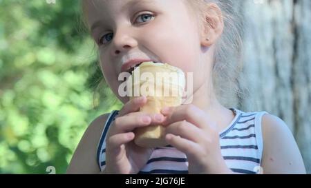 Nettes kleines Mädchen isst draußen Eis. Nahaufnahme eines blonden Mädchens, das auf der Parkbank sitzt und Eis isst. Odessa, Ukraine Stockfoto