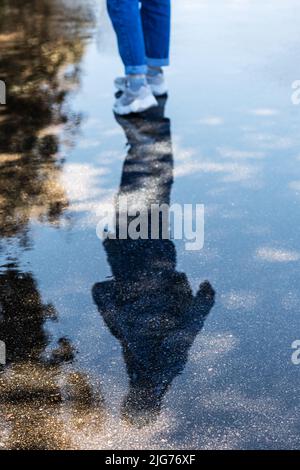 Die Silhouette einer wandelenden Frau spiegelt sich in einer Pfütze auf nassem Bürgersteig wider. Lauf dem Regen nach. Selektiver Fokus. Stockfoto